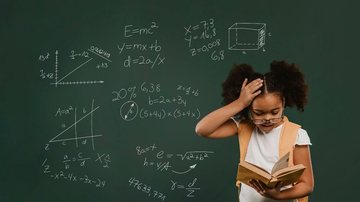 Menina segurando livro na frente de um quadro negro com contas matemáticas.
