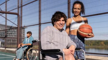 Homens na cadeira de roda e uma mulher de pé segurando uma bola de basquete em uma quadra