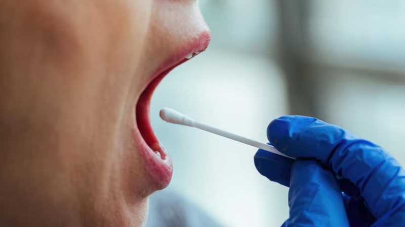 Mulher realizando teste de saliva com a boca aberta e profissional da saúde segurando haste com algodão