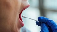 Mulher realizando teste de saliva com a boca aberta e profissional da saúde segurando haste com algodão