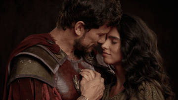 casal trajando roupas de época, com os rostos próximos, sob fundo escuro.