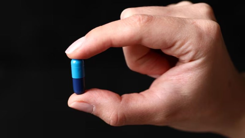 mão segurando cápsula de medicamento azul sob fundo preto.