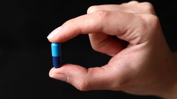 mão segurando cápsula de medicamento azul sob fundo preto.
