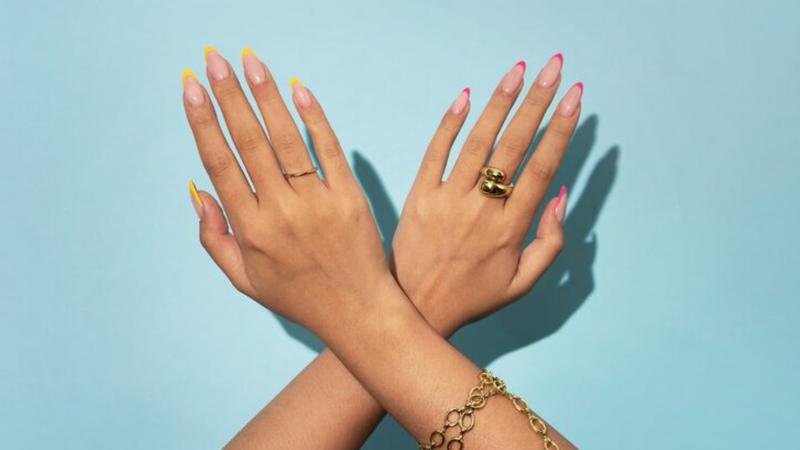 Unhas no estilo francesinha em cores distintas, dispostas sob fundo azul neutro.