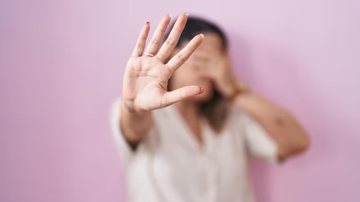 Mulher cobrindo proprio rosto e apontando com a mão aberta em direção à câmera