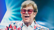 Elton John posando de forma animada e sorridente sob fundo com luzes azuis.