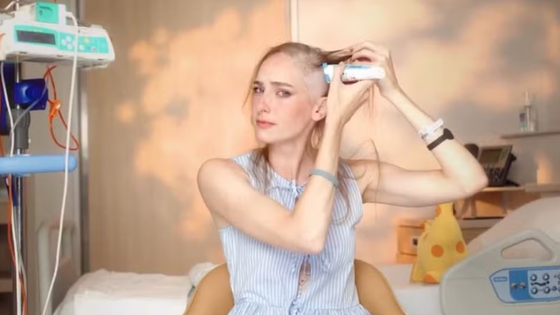 Atriz Dani Gondim trajando azul e raspando os cabelos sob fundo de quarto de hospital