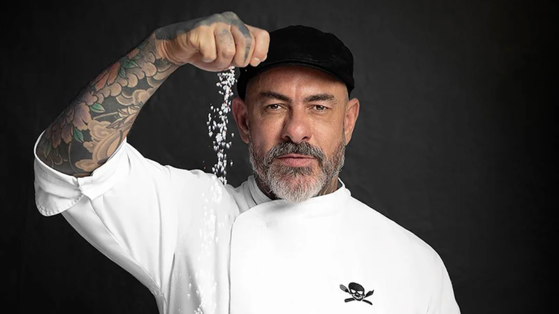 Chef Henrique Fogaça posando sob fundo neutro e despejando sal com a ponta dos dedos