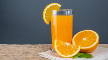 copo de suco de laranja ao lado de laranjas cortadas sob fundo neutro.