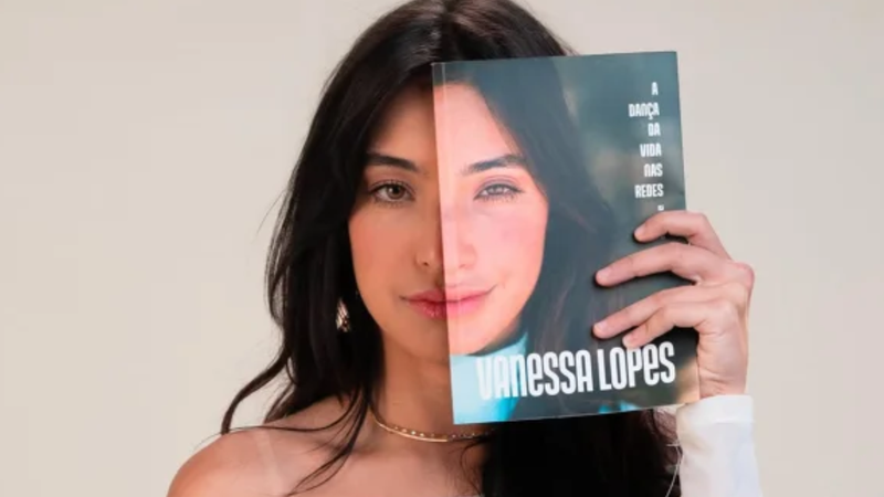Vanessa Lopes, posando sob fundo neutro, segurando o seu livro em frente ao seu rosto.