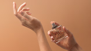 mão segurando perfume, aplicando fragrância sob fundo de cor coral