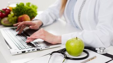 Nutricionista usando o computador, com estetoscópio ao lado e maçã verde ao outro.