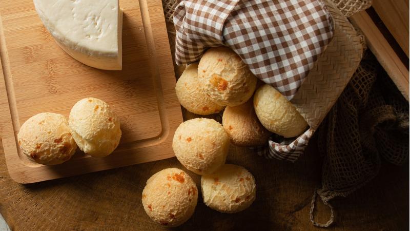 diversas unidades de pão de queijo em uma tábua de madeira com um pedaço de queijo minas ao fundo